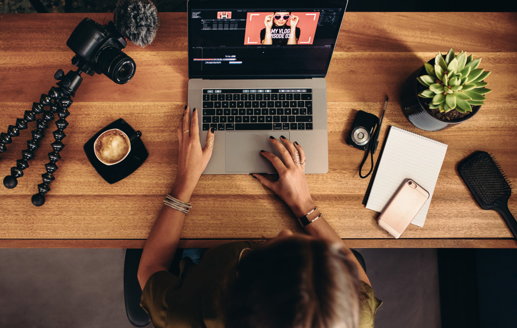 High angle view of female vlogger editing video on laptop. Young woman working on computer with cameras and accessories on table.