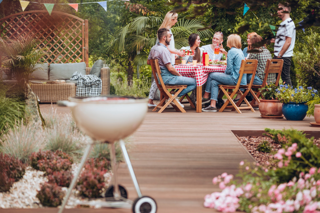 Wooden patio in the garden with a grill standing in the front and happy young people gathered around a table full of food during summer brake meeting