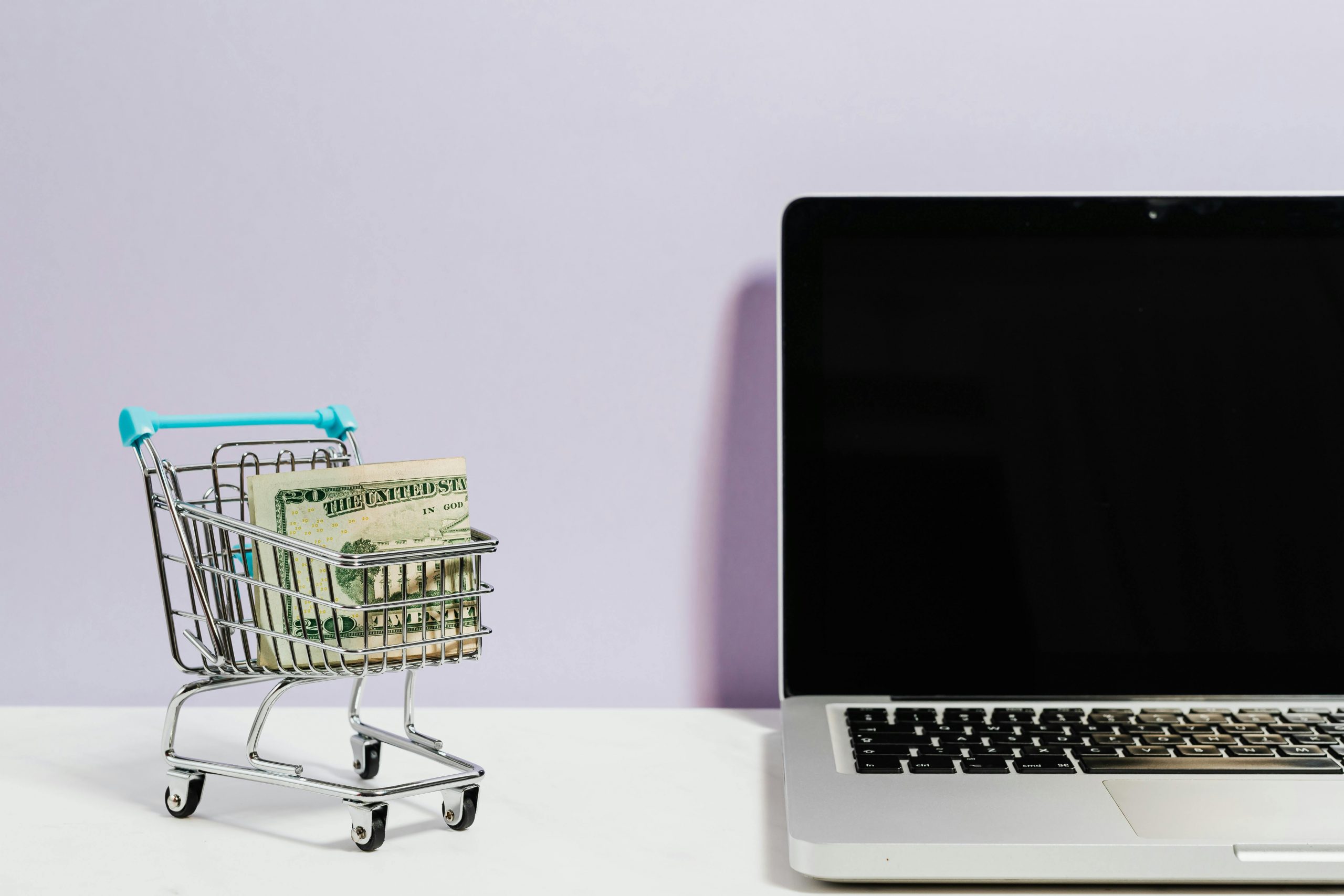 miniature shopping cart filled with money next to a laptop