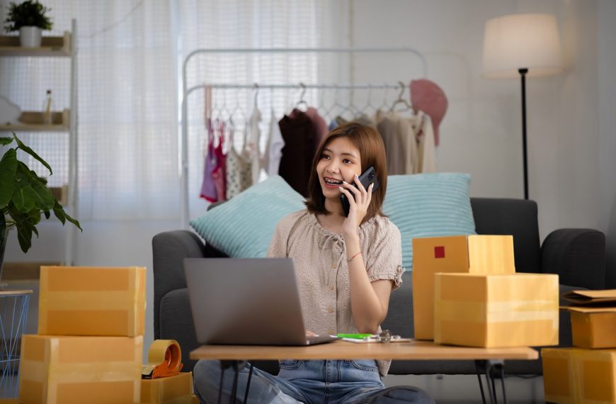 woman talking on her phone at a desk with a laptop and cardboard boxes